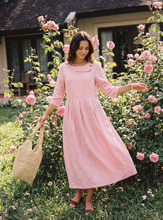 Linen maxi dress with crescent neck, decorative buttons, and pleated waist in soft pink from LINTICO, styled for a flowing, elegant look.